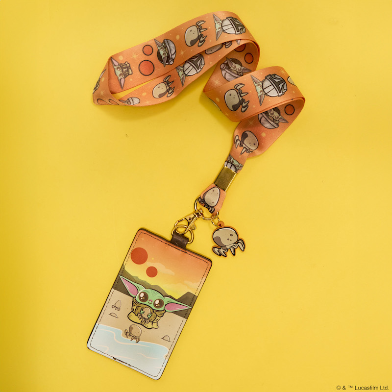Orange lanyard featuring images of Grogu, Mando, and crab-like creatures in a repeating patten. Attached is a card holder, featuring Grogu standing on a beach holding a crab-like creature. Two more surround him on the beach. Another crab-like creature appears on the lanyard as a silicone charm. The lanyard with card holder lays against a yellow background. 